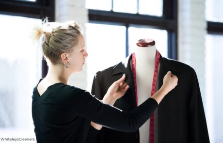 A focused dry cleaner, wearing a white lab coat and a blue undershirt, meticulously inspects a light-colored garment hanging in a well-lit dry cleaning shop. Rows of neatly organized clothes, some protected by plastic coverings, are visible in the background, suggesting a professional and clean environment at Whiteleys Dry Cleaning in West Wickham.